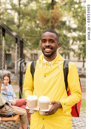 Happy young contemporary male courier in yellow jacket holding two drinks 68954969