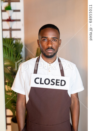 Young serious waiter of African ethnicity with paper saying closed on his neck Young serious waiter of African ethnicity with paper saying closed on his neck 68955011