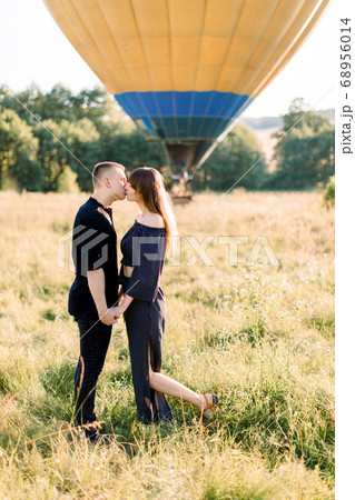 Cute Caucasian young couple in love, standing in summer field, holding hands and kissing. Hot colorful air balloon at sunrise on the background. Anniversary or engagement celebration Cute Caucasian young couple in love, standing in summer field, holding hands and kissing. Hot colorful air balloon at sunrise on the background. Anniversary or engagement celebration 68956014