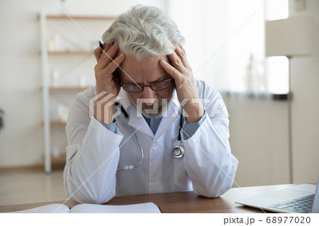 Exhausted unhappy mature doctor wearing glasses sitting at desk Exhausted unhappy mature doctor wearing glasses sitting at desk 68977020