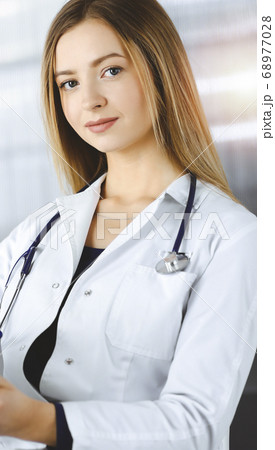 Young optimistic woman-doctor is using a clipboard, while sitting at the desk in her sunny cabinet in a clinic. Portrait of friendly female physician woman with a stethoscope. Perfect medical service 68977028