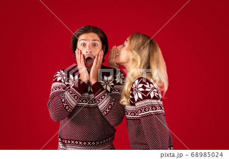 Shocked millennial guy in Christmas outfit and girl whispering something into his ear on red background Shocked millennial guy in Christmas outfit and girl whispering something into his ear on red background 68985024