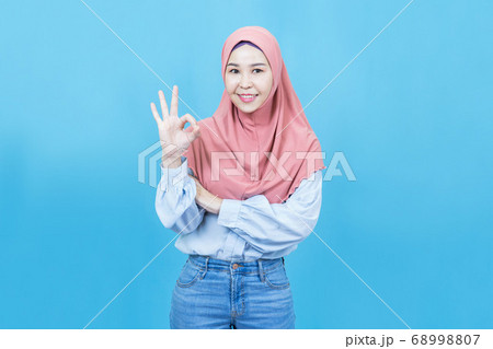 Portrait of young beautiful asian muslim woman in traditional dress make gesture Okay isolated on blue background Portrait of young beautiful asian muslim woman in traditional dress make gesture Okay isolated on blue background 68998807