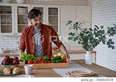 Vegetarianism concept. Happy young man in casual clothes holding basket with various fresh vegetables and smiling, cooking healthy food at home 69002678