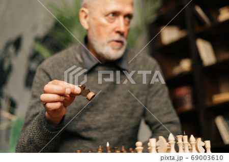 Caucasian mature adult bearded gray-haired man making move at chess game, focused on foreground. Caucasian mature adult bearded gray-haired man making move at chess game, focused on foreground. 69003610
