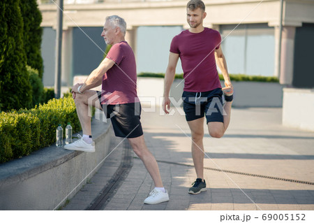 Grey-haired man stretching knee, young male stretching quad next to him 69005152