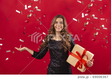Sexy young woman in evening dress holding gift box and posing over red background with falling confetti Sexy young woman in evening dress holding gift box and posing over red background with falling confetti 69006367
