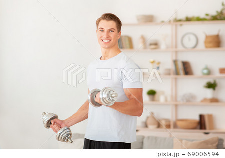 Strength training at home during quarantine. Smiling handsome guy lifts dumbbells Strength training at home during quarantine. Smiling handsome guy lifts dumbbells 69006594