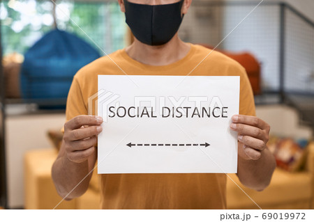 Close up shot of office worker wearing black protective mask showing paper with text SOCIAL DISTANCE at camera while standing in the office Close up shot of office worker wearing black protective mask showing paper with text SOCIAL DISTANCE at camera while standing in the office 69019972