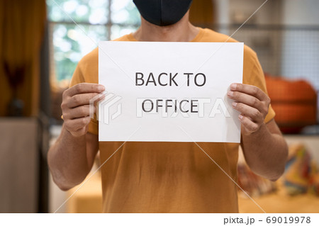 Cropped shot of a male office worker wearing black protective mask showing paper with text BACK TO OFFICE at camera Business after lockdown 69019978