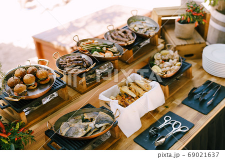 Buffet table with copper pans and prepared dishes for the celebration. Fried fish in a pan, mini burgers, focaccia and Serbian sausages - cevapcici, and grilled squid. Buffet table with copper pans and prepared dishes for the celebration. Fried fish in a pan, mini burgers, focaccia and Serbian sausages - cevapcici, and grilled squid. 69021637