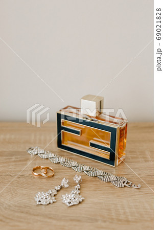 A perfume bottle, a bracelet, earrings-studs and wedding rings on a wooden table against a light wall. 69021828