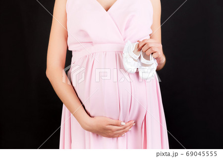 Cropped image of pregnant woman in pink dress holding baby girl booties against her belly at black background. Happy pregnancy. Copy space Cropped image of pregnant woman in pink dress holding baby girl booties against her belly at black background. Happy pregnancy. Copy space 69045555