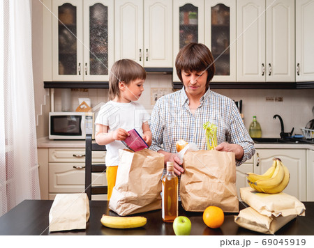 Family sorts out purchases in the kitchen. Father and son tastes products in bags made of craft paper. Food delivery in conditions of quarantine because of coronavirus COVID19. 69045919