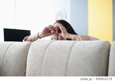 Woman looks out from behind sofa and holds her hands through binoculars. Woman looks out from behind sofa and holds her hands through binoculars. 69064229