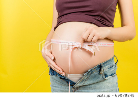 Close up of a pregnant woman in opened jeans measuring her belly with a tape measure at colorful background with copy space. entimeter measurement Close up of a pregnant woman in opened jeans measuring her belly with a tape measure at colorful background with copy space. entimeter measurement 69079849
