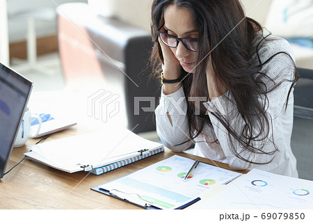 Sad woman sits at her work desk in office over charts with business indicators and holds her head. Sad woman sits at her work desk in office over charts with business indicators and holds her head. 69079850