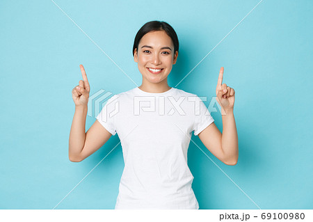 Gorgeous smiling young 20s female asian in casual clothes, pointing fingers up, showing advertisement, demonstrating promo offer. Korean girl indicating at banner, blue background 69100980