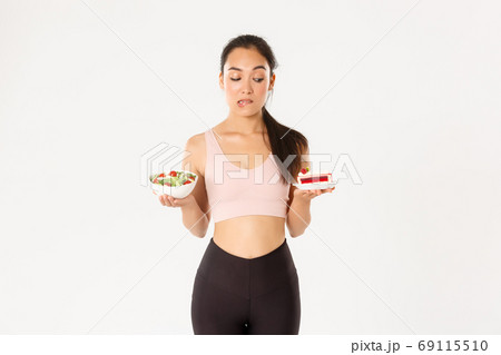 Active lifestyle, fitness and wellbeing concept. Portrait of indecisive and tempting cute asian girl trying resist temptation as looking at delicious cake, being on diet, need eat healthy salad 69115510
