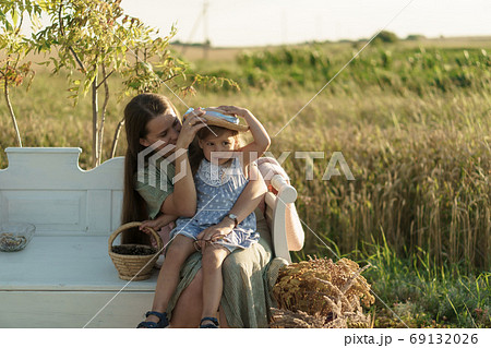childhood, motherhood, provence style concept - happy small 3 year old blonde little girl in blue dress slavic appearance sits on white bench in braided hat with mom on field on summer before sunset. 69132026