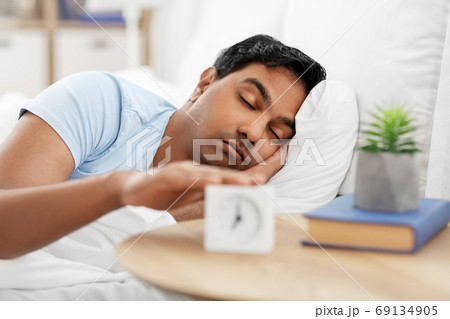 sleepy indian man with alarm clock lying in bed 69134905