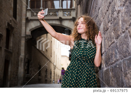 Cheerful young girl standing near brick wall and making selfie with phone 69137975