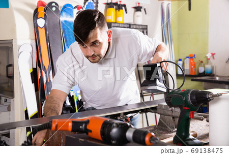 Confident master of ski service professionally performing sharpening of edges on skis Confident master of ski service professionally performing sharpening of edges on skis 69138475