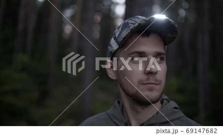 Portrait of handsome young man hunter or tourist. Man puts on his head cap with diode flashlight on visor in evening. Portrait of handsome young man hunter or tourist. Man puts on his head cap with diode flashlight on visor in evening. 69147682