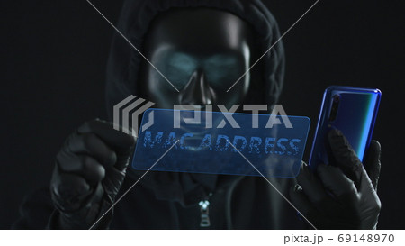 Hacker wearing black mask pulls MAC ADDRESS tab from a smartphone. Hacking concept Hacker wearing black mask pulls MAC ADDRESS tab from a smartphone. Hacking concept 69148970