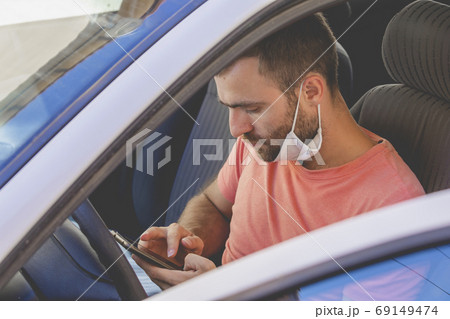 A young man, wearing a cloth face mask, checks the mobile phone inside the car 69149474