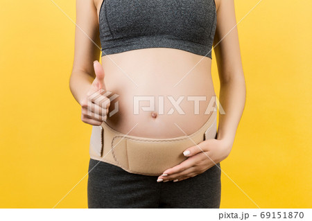 Close up of pregnant woman showing thumb up and wearing elastic pregnancy bandage to make the pain go away at yellow background with copy space. Orthopedic abdominal support belt concept Close up of pregnant woman showing thumb up and wearing elastic pregnancy bandage to make the pain go away at yellow background with copy space. Orthopedic abdominal support belt concept 69151870