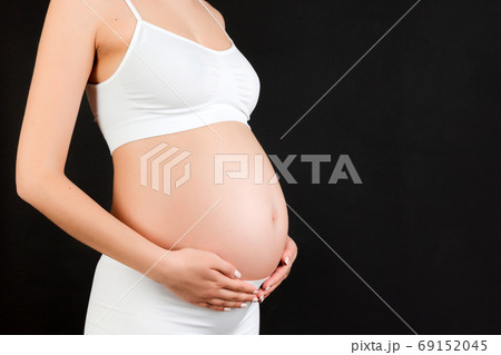 Cropped image of pregnant woman in white underwear embracing her big abdomen at black background. Maternity concept. Copy space Cropped image of pregnant woman in white underwear embracing her big abdomen at black background. Maternity concept. Copy space 69152045