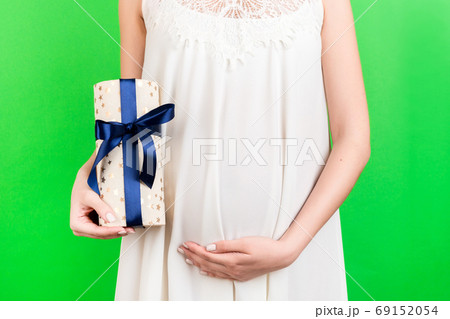 Cropped image of pregnant woman in white dress holding a gift box and touching her belly at green background. Expecting a baby boy. Copy space Cropped image of pregnant woman in white dress holding a gift box and touching her belly at green background. Expecting a baby boy. Copy space 69152054