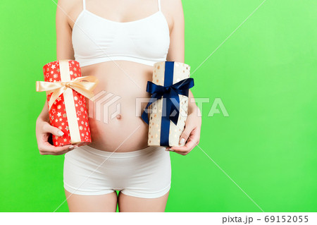 Close up of two gift boxes in pregnant woman's hands against her belly at green background. Is it a boy or a girl? Waiting for twins. Copy space 69152055