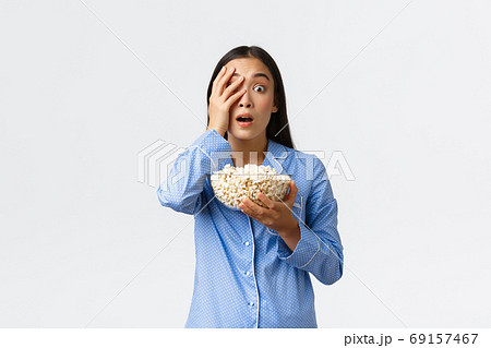 Home leisure, sleepover and slumber party concept. Shocked asian girl in pajamas holding popcorn, gasping and looking startled, open mouth with interest as watching tv or movie, white background 69157467
