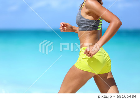 Female runner on beach with sports bra and shorts. Midsection closeup of body of a woman athlete running with speed fast training cardio wearing neon yellow activewear outfit. Active lifestyle. Female runner on beach with sports bra and shorts. Midsection closeup of body of a woman athlete running with speed fast training cardio wearing neon yellow activewear outfit. Active lifestyle. 69165484