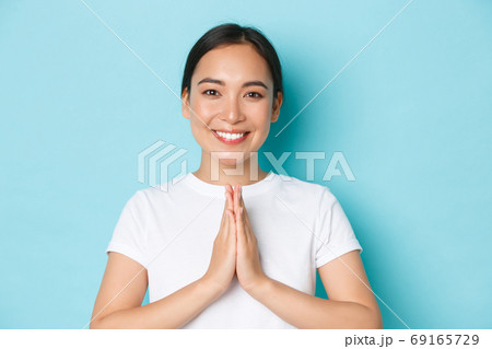 Close-up of pleasant attractive asian girl smiling, holding hands in plead, praying gesture, thanking for something, show namaste sign, standing over light blue background 69165729