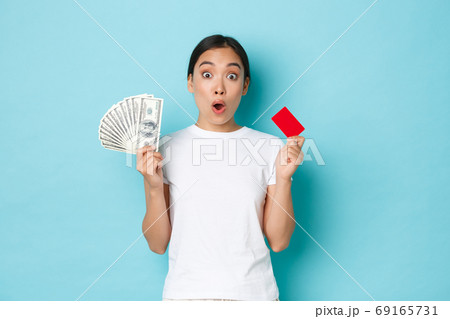 Shopping, money and finance concept. Amused asian girl in white casual t-shirt gasping, found out awesome prices, discount offers in shop, holding both credit card and cash, light-blue background Shopping, money and finance concept. Amused asian girl in white casual t-shirt gasping, found out awesome prices, discount offers in shop, holding both credit card and cash, light-blue background 69165731