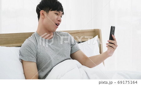 Young happy man using mobile phone for video call in bed at home 69175164