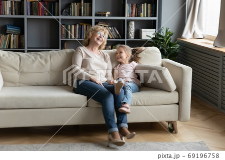 Overjoyed little granddaughter and mature grandmother laughing, sitting on couch 69196758
