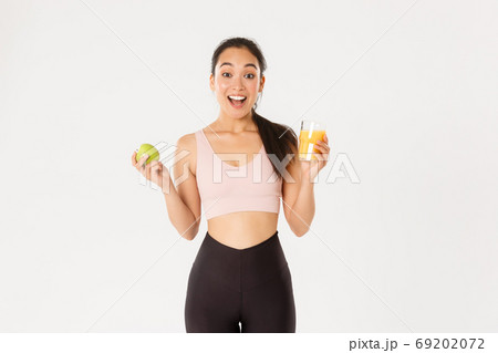 Sport, wellbeing and active lifestyle concept. Excited cute asian fitness girl, sportswoman with apple and orange juice gasping amazed and happy, eating healthy to stay fit, white background 69202072