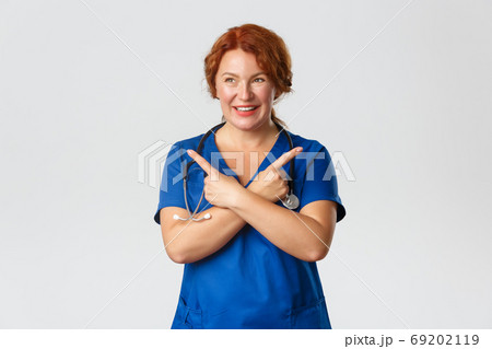Medicine, healthcare and coronavirus concept. Cheerful smiling middle-aged redhead doctor, physician or nurse in blue scrubs showing way, medical worker pointing sideways, choosing 69202119