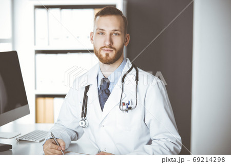 Friendly red-bearded doctor sitting and writing at clipboard in clinic. Perfect medical service in hospital. Medicine concept Friendly red-bearded doctor sitting and writing at clipboard in clinic. Perfect medical service in hospital. Medicine concept 69214298