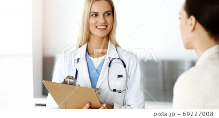Woman-doctor at work in sunny hospital is happy to consult female patient. Blonde physician checks medical history record and exam results while using clipboard Woman-doctor at work in sunny hospital is happy to consult female patient. Blonde physician checks medical history record and exam results while using clipboard 69227008