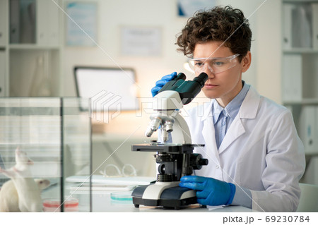 Contemporary young female researcher looking at antibodies in microscope 69230784