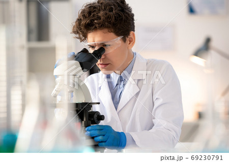 Young serious scientist or lab worker bending over microscope by workplace 69230791