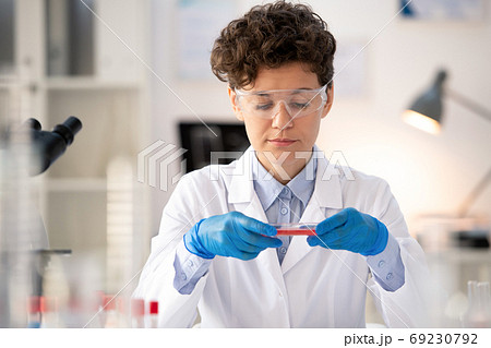Contemporary female scientist in whitecoat and gloves sitting by workplace 69230792