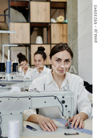 Young pretty seamstress in workwear looking at you while repairing clothes Young pretty seamstress in workwear looking at you while repairing clothes 69230945