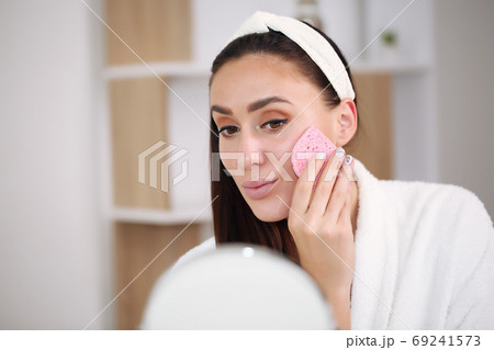 brunette in a bathrobe washes off makeup with a pink sponge 69241573