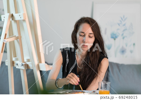 Girl artist, at home on sofa, background wall painting easel, brush and color palette with paints. Begins to paint. Black T-shirt long hair. Wooden stand for a picture. Girl artist, at home on sofa, background wall painting easel, brush and color palette with paints. Begins to paint. Black T-shirt long hair. Wooden stand for a picture. 69248015